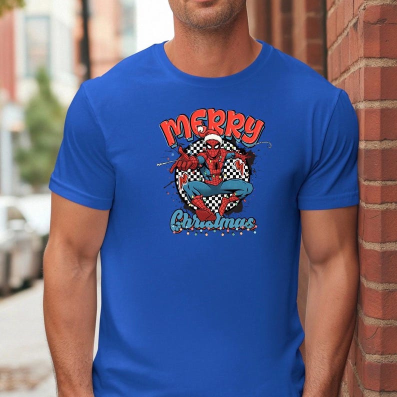 Puede incluir: Camiseta azul con un dise&ntilde;o de Spider-Man con un gorro de Pap&aacute; Noel y las palabras "Merry Christmas" en rojo y azul. El superh&eacute;roe est&aacute; sobre un fondo a cuadros con salpicaduras de pintura.