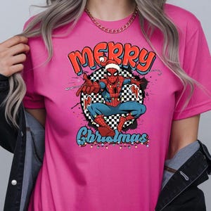 Puede incluir: Una camiseta rosa con un dise&ntilde;o de Spider-Man con un gorro de Pap&aacute; Noel, las palabras "Merry Christmas", un fondo a cuadros y luces navide&ntilde;as. El dise&ntilde;o tiene un tema festivo navide&ntilde;o.