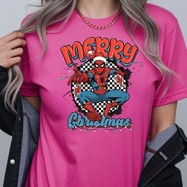 Puede incluir: Una camiseta rosa con un dise&ntilde;o de Spider-Man con un gorro de Pap&aacute; Noel, las palabras "Merry Christmas", un fondo a cuadros y luces navide&ntilde;as. El dise&ntilde;o tiene un tema festivo navide&ntilde;o.