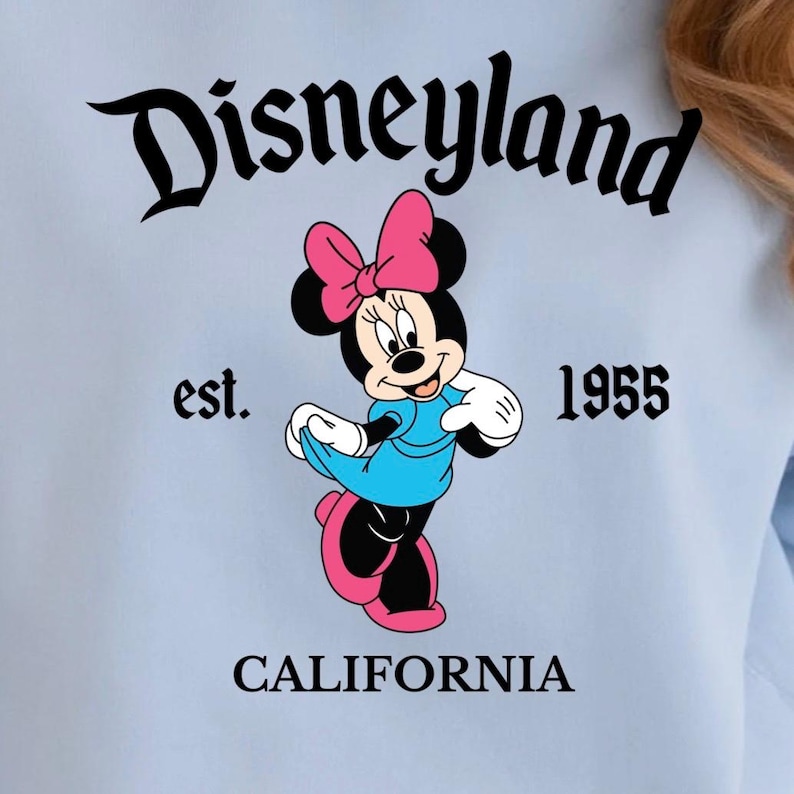Puede incluir: Gr&aacute;fico azul claro con Minnie Mouse con vestido azul y lazo rosa, con el texto "Disneyland" arriba y "est. 1955 CALIFORNIA" abajo. La ilustraci&oacute;n es de estilo vintage.