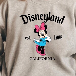 Puede incluir: Una sudadera gris claro con un gr&aacute;fico de Minnie Mouse. El gr&aacute;fico incluye la palabra "Disneyland", el texto "est. 1955 CALIFORNIA" y una Minnie Mouse de dibujos animados con un vestido azul y un lazo rosa.