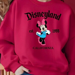 Puede incluir: Una sudadera roja con el logotipo de Disneyland y un dibujo de Minnie Mouse con un vestido azul y un lazo rosa. Tambi&eacute;n se muestra el texto "est. 1955" y "CALIFORNIA". La sudadera es una prenda vibrante y llamativa.