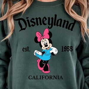 Puede incluir: Sudadera verde con la palabra "Disneyland" en escritura negra sobre un dibujo animado de Minnie Mouse con un vestido azul y un lazo rosa. El texto "est. 1955 CALIFORNIA" tambi&eacute;n est&aacute; impreso en la sudadera.