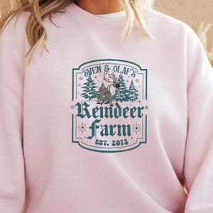 May include: A light pink sweatshirt featuring a teal graphic that reads "Sven & Olaf's Reindeer Farm Est. 2013". The graphic includes a reindeer, pine trees, and decorative elements.
