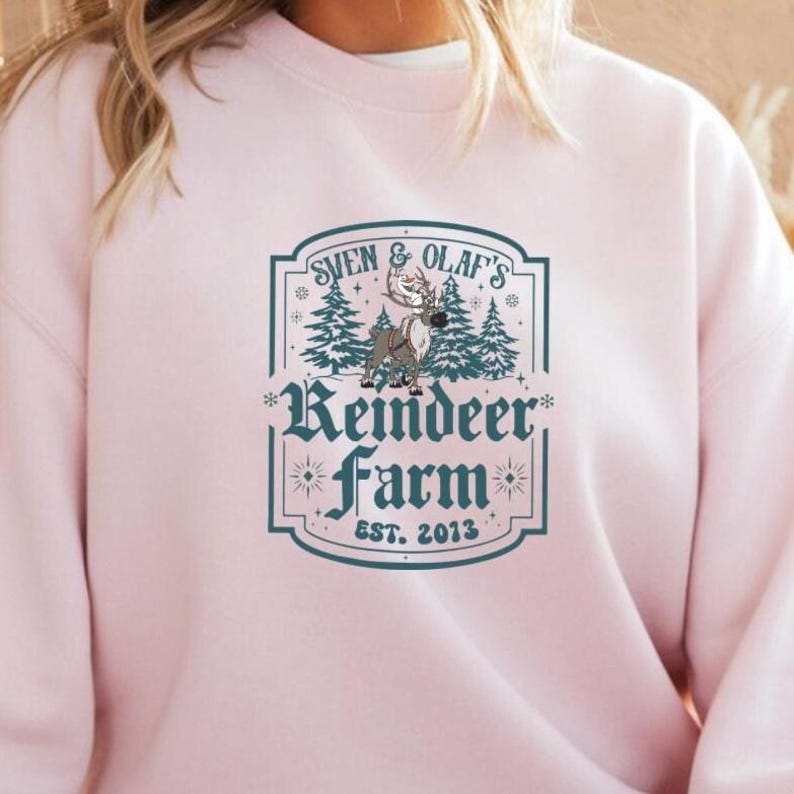 May include: A light pink sweatshirt featuring a teal graphic that reads "Sven & Olaf's Reindeer Farm Est. 2013". The graphic includes a reindeer, pine trees, and decorative elements.