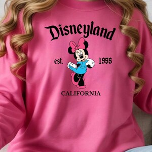 Puede incluir: Sudadera rosa con el logotipo de Disneyland y un gr&aacute;fico de Minnie Mouse. El gr&aacute;fico incluye el texto "est. 1955" y "CALIFORNIA". Minnie Mouse lleva un vestido azul y un lazo rosa.