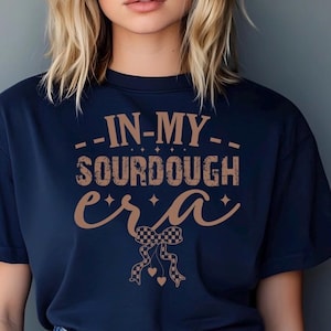 May include: Navy blue t-shirt with the text "--IN-MY-- SOURDOUGH era" in brown, featuring a checkered bow and hearts. The shirt is a casual wear item, perfect for those who enjoy baking sourdough bread.