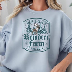 May include: A light blue sweatshirt with a graphic that reads "Sven & Olaf's Reindeer Farm EST. 2013." The graphic features a reindeer, pine trees, and decorative elements in shades of green and brown.