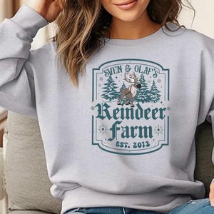 May include: A light gray sweatshirt featuring a teal and white graphic. The graphic reads "Sven & Olaf's Reindeer Farm EST. 2013" with an illustration of a reindeer and pine trees. The sweatshirt has long sleeves.