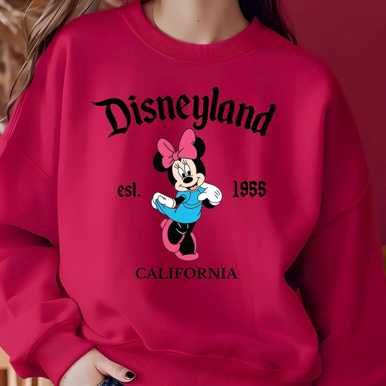 Puede incluir: Una sudadera roja con la palabra "Disneyland" en letra cursiva, con "est. 1955" y "CALIFORNIA" debajo. Un gr&aacute;fico de Minnie Mouse est&aacute; en el centro, con un vestido azul y un lazo rosa. Un tema cl&aacute;sico de Disney.