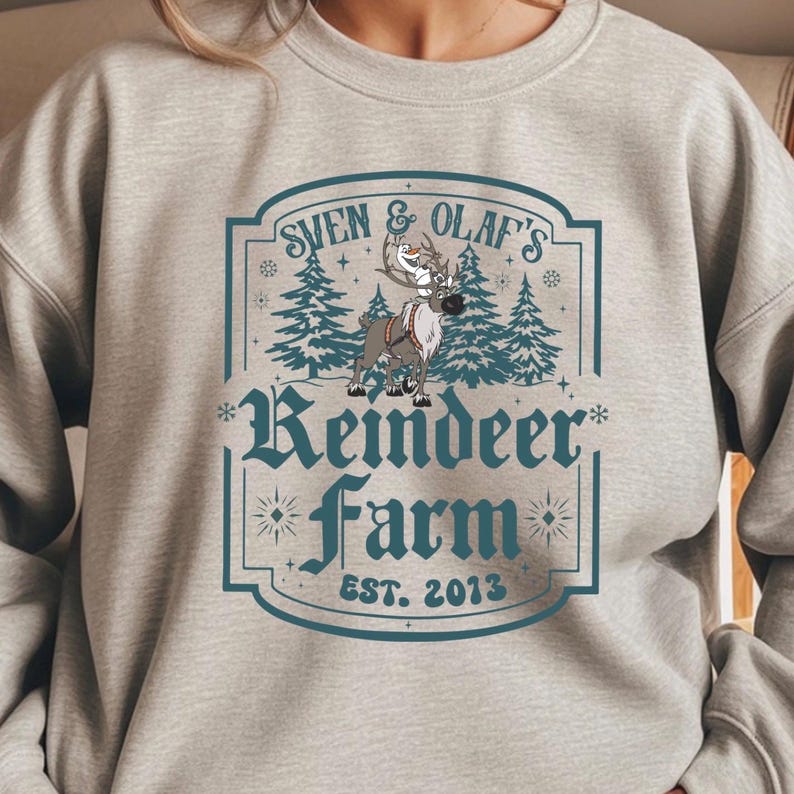 May include: A light gray sweatshirt featuring a teal and white graphic. The graphic reads "Sven & Olaf's Reindeer Farm EST. 2013" with illustrations of reindeer and pine trees. The sweatshirt is a crew neck style.