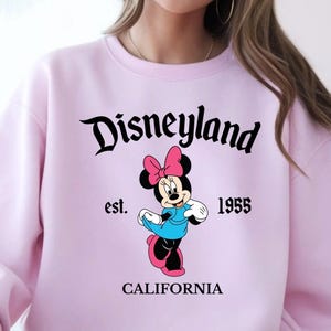 Puede incluir: Una sudadera rosa claro con la palabra "Disneyland" en escritura negra sobre un gr&aacute;fico de Minnie Mouse. El gr&aacute;fico incluye el texto "est. 1955 CALIFORNIA". Minnie Mouse lleva un vestido azul y un lazo rosa.