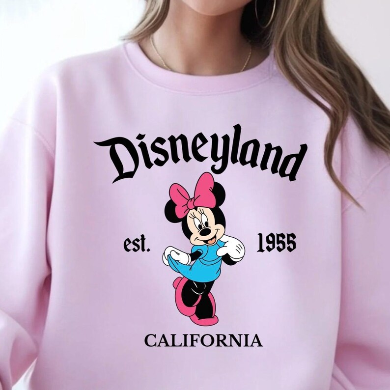 Puede incluir: Una sudadera rosa claro con la palabra "Disneyland" en escritura negra sobre un gr&aacute;fico de Minnie Mouse. El gr&aacute;fico incluye el texto "est. 1955 CALIFORNIA". Minnie Mouse lleva un vestido azul y un lazo rosa.