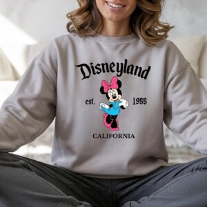 Puede incluir: Sudadera gris claro con el logotipo de Disneyland y Minnie Mouse. El dise&ntilde;o incluye el texto "Disneyland est. 1955 CALIFORNIA". Minnie Mouse lleva un lazo rosa, un vestido azul y zapatos rosas.