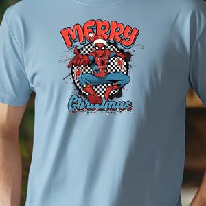 Puede incluir: Camiseta azul claro con un dise&ntilde;o de Spider-Man con gorro de Pap&aacute; Noel y la frase "Merry Christmas". El dise&ntilde;o incluye un tablero de ajedrez y luces navide&ntilde;as.