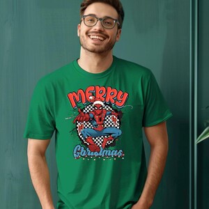 Puede incluir: Camiseta verde con un gr&aacute;fico de Spider-Man con un gorro de Pap&aacute; Noel y las palabras "MERRY Christmas" en rojo y azul. El superh&eacute;roe est&aacute; sobre un fondo a cuadros en blanco y negro.