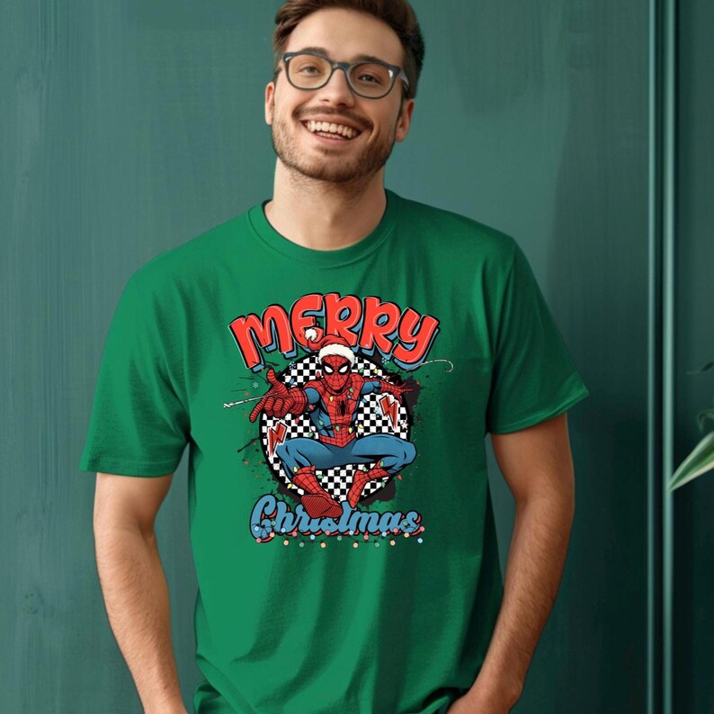 Puede incluir: Camiseta verde con un gr&aacute;fico de Spider-Man con un gorro de Pap&aacute; Noel y las palabras "MERRY Christmas" en rojo y azul. El superh&eacute;roe est&aacute; sobre un fondo a cuadros en blanco y negro.