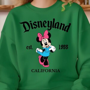 Puede incluir: Sudadera verde con el logotipo de Disneyland en escritura negra, y Minnie Mouse con un vestido azul y un lazo rosa. Tambi&eacute;n se muestra el texto "est. 1955 CALIFORNIA". Una prenda de vestir cl&aacute;sica con tem&aacute;tica de Disney.