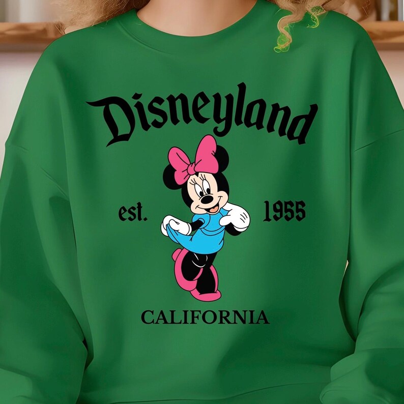 Puede incluir: Sudadera verde con el logotipo de Disneyland en escritura negra, y Minnie Mouse con un vestido azul y un lazo rosa. Tambi&eacute;n se muestra el texto "est. 1955 CALIFORNIA". Una prenda de vestir cl&aacute;sica con tem&aacute;tica de Disney.