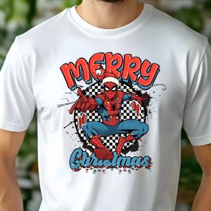 Puede incluir: Camiseta blanca con un dise&ntilde;o de Spider-Man con gorro de Pap&aacute; Noel, rodeado de luces navide&ntilde;as y las palabras "Merry Christmas". El dise&ntilde;o incluye un fondo a cuadros y detalles coloridos.