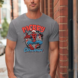 Puede incluir: Camiseta gris con un gr&aacute;fico de Spider-Man con un gorro de Pap&aacute; Noel y las palabras "Merry Christmas" en rojo y azul. El superh&eacute;roe est&aacute; sobre un fondo a cuadros blanco y negro, con luces navide&ntilde;as debajo.