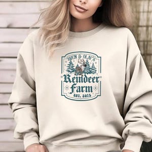 May include: Cream-colored sweatshirt featuring a graphic with the text "Sven & Olaf's Reindeer Farm EST. 2013". The graphic includes a reindeer, pine trees, and decorative elements in shades of blue and brown.