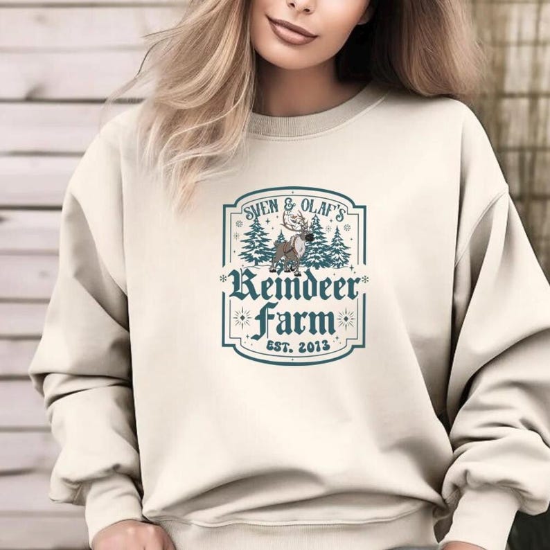 May include: Cream-colored sweatshirt featuring a graphic with the text "Sven & Olaf's Reindeer Farm EST. 2013". The graphic includes a reindeer, pine trees, and decorative elements in shades of blue and brown.