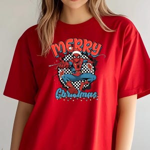 Puede incluir: Camiseta roja con un dise&ntilde;o de Spider-Man con gorro de Pap&aacute; Noel. El dise&ntilde;o incluye las palabras "Merry Christmas" en rojo y azul, sobre un fondo a cuadros blanco y negro.