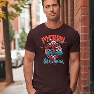 Puede incluir: Camiseta granate con un gr&aacute;fico de Spider-Man con un gorro de Pap&aacute; Noel y las palabras "Merry Christmas". El gr&aacute;fico de Spider-Man est&aacute; sobre un fondo a cuadros en blanco y negro.