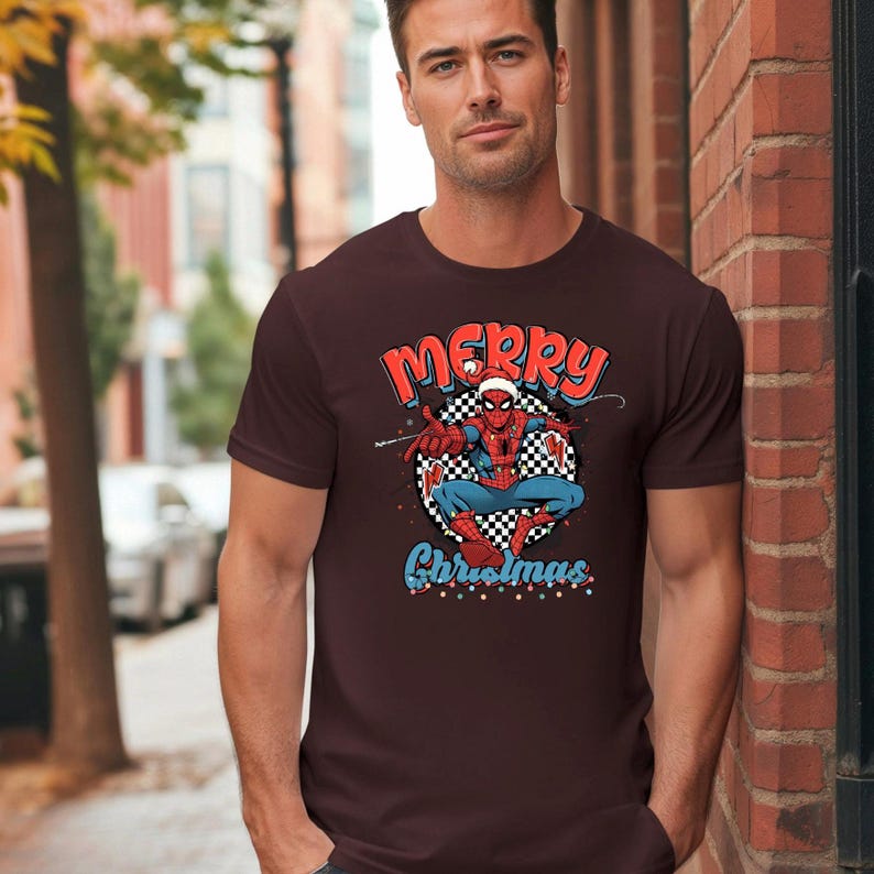 Puede incluir: Camiseta granate con un gr&aacute;fico de Spider-Man con un gorro de Pap&aacute; Noel y las palabras "Merry Christmas". El gr&aacute;fico de Spider-Man est&aacute; sobre un fondo a cuadros en blanco y negro.