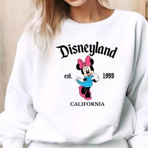 Puede incluir: Sudadera blanca con el logotipo de Disneyland en escritura negra y un gr&aacute;fico de Minnie Mouse. Minnie lleva un lazo rosa, un vestido azul y zapatos rosas. El texto "est. 1955 CALIFORNIA" est&aacute; impreso debajo.