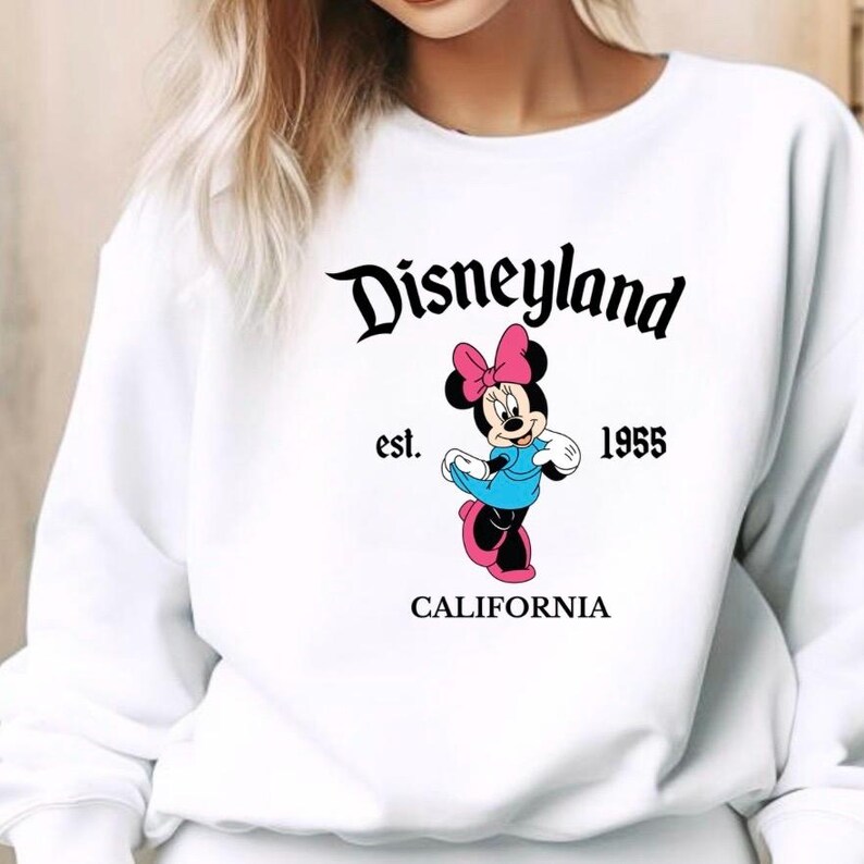 Puede incluir: Sudadera blanca con el logotipo de Disneyland en escritura negra y un gr&aacute;fico de Minnie Mouse. Minnie lleva un lazo rosa, un vestido azul y zapatos rosas. El texto "est. 1955 CALIFORNIA" est&aacute; impreso debajo.