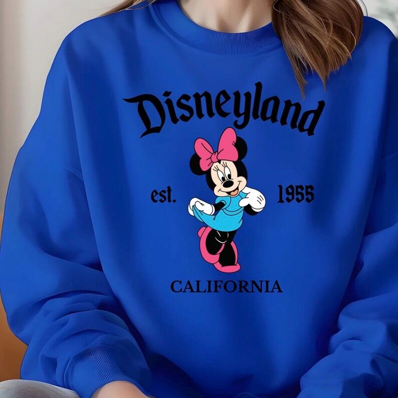 Puede incluir: Sudadera azul real con un gr&aacute;fico de Minnie Mouse con lazo, vestido y zapatos rosas. La palabra "Disneyland" est&aacute; arqueada en la parte superior en negro, con "est. 1955" y "CALIFORNIA" debajo.