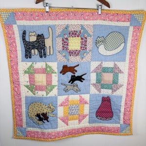 May include: A handmade quilt with a patchwork design. It features blue squares with appliqued cat figures. The quilt has a pink floral border and various patterned fabrics in the squares, including floral and geometric designs. It is hanging on a wooden hanger.
