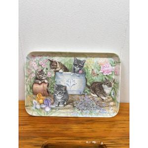 Puede incluir: Bandeja rectangular decorativa con una ilustración de cinco gatitos en un jardín. Los gatitos están rodeados de flores, una maceta azul y una mariposa. La bandeja tiene un borde blanco y una superficie de madera.