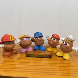Pode incluir: Cinco figuras do Mr. Potato Head em várias roupas. As figuras são marrons com chapéus e roupas de várias cores, incluindo um bombeiro, um médico e um policial. Os brinquedos estão em uma superfície de madeira.