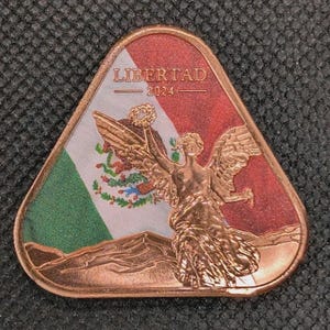 May include: A copper-colored triangular coin featuring the Mexican flag and a depiction of the Angel of Independence. The coin has the word "LIBERTAD" and the year "2024" printed on it.