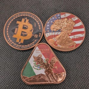 May include: Three copper coins: a Bitcoin design, a walking Liberty design with an American flag background, and a Mexican Libertad coin. The Bitcoin coin has the text "ONE AVDP OUNCE" and "999 FINE COPPER". The Libertad coin has the text "LIBERTAD 2024".