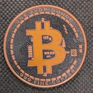 May include: A copper Bitcoin coin with a black and copper design. The coin features the Bitcoin symbol in the centre, surrounded by the text "ONE AVDP OUNCE" and "999 FINE COPPER". The coin has a detailed, textured surface.