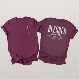 May include: Two maroon t-shirts. One shirt features a white cross on the front. The other shirt displays the word "BLESSED" in white, along with "John 20:29" and a Bible verse.