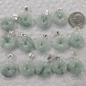 Puede incluir: Una colección de colgantes de jade en forma de rosquilla de color verde pálido con colgantes plateados. Los colgantes tienen forma circular con un agujero en el centro. Se incluye una moneda para comparar el tamaño.