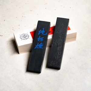 Op de afbeelding: Twee zwarte inktstokken, een met Chinese karakters in blauwe inkt die "Plaats van Vuur" lezen en de andere met een gesneden ontwerp van een dennenboom en een vogel. De inktstokken liggen op een houten doos met een rood en wit label.