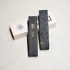 Op de afbeelding: Twee zwarte inktstokken in houten dozen. De inktstokken hebben gouden letters en ontwerpen. Een inktstok heeft de tekst "Black King" in gouden letters. De andere inktstok heeft een ontwerp van een boom met een gouden zegel.