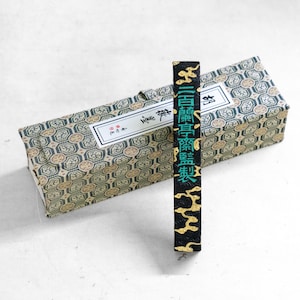 Op de afbeelding: Een zwarte inktstaaf met gouden accenten en groene tekst, geplaatst voor een doos met patronen. De doos en de inktstaaf hebben Aziatisch geïnspireerde ontwerpen. De tekst op de inktstaaf is in het Japans.