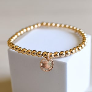 Peut inclure: Bracelet de perles dorées avec un petit charme rond. Le charme porte l'inscription "YOU TRUST JESUS". Le bracelet est composé de petites perles rondes de couleur or. Le bracelet est présenté sur une surface blanche.