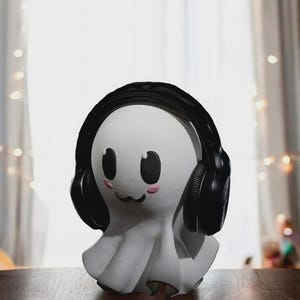 May include: A white ghost-shaped figurine with black headphones. The ghost has black eyes, a small black mouth, and pink cheeks. The figurine is sitting on a wooden surface, with a blurred background of a window and string lights.