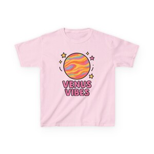 Pode incluir: Camiseta rosa com um planeta de desenho animado e estrelas. O texto "Venus Vibes" é impresso em rosa e preto.
