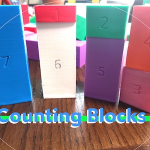 May include: Counting blocks in different colors and sizes, stacked on top of each other. The blocks are labeled with numbers 1 through 7. The blocks are on a wooden surface.