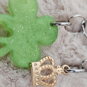 May include: A green, glitter-flecked shamrock charm with a gold-colored crown charm attached to a silver-colored chain. The shamrock has a heart-shaped cutout. The chain is made of interlocking oval links.