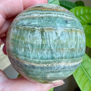 May include: A polished spherical stone featuring horizontal bands of sage green, forest green, and translucent white. The surface displays natural mineral inclusions, crystalline textures, and thin golden-brown layers throughout the round object.